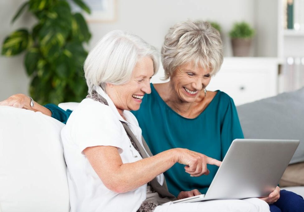 Two senior women surfing the internet laughing and exclaiming as they point to something on the screen while sitting side by side on a sofa in the house